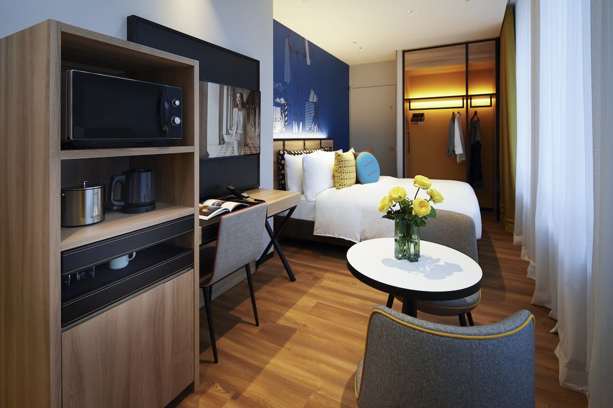 A modern hotel room with a wooden desk, a microwave, a coffee maker, a bed with white bedding and yellow flowers, and a small round table with a vase.