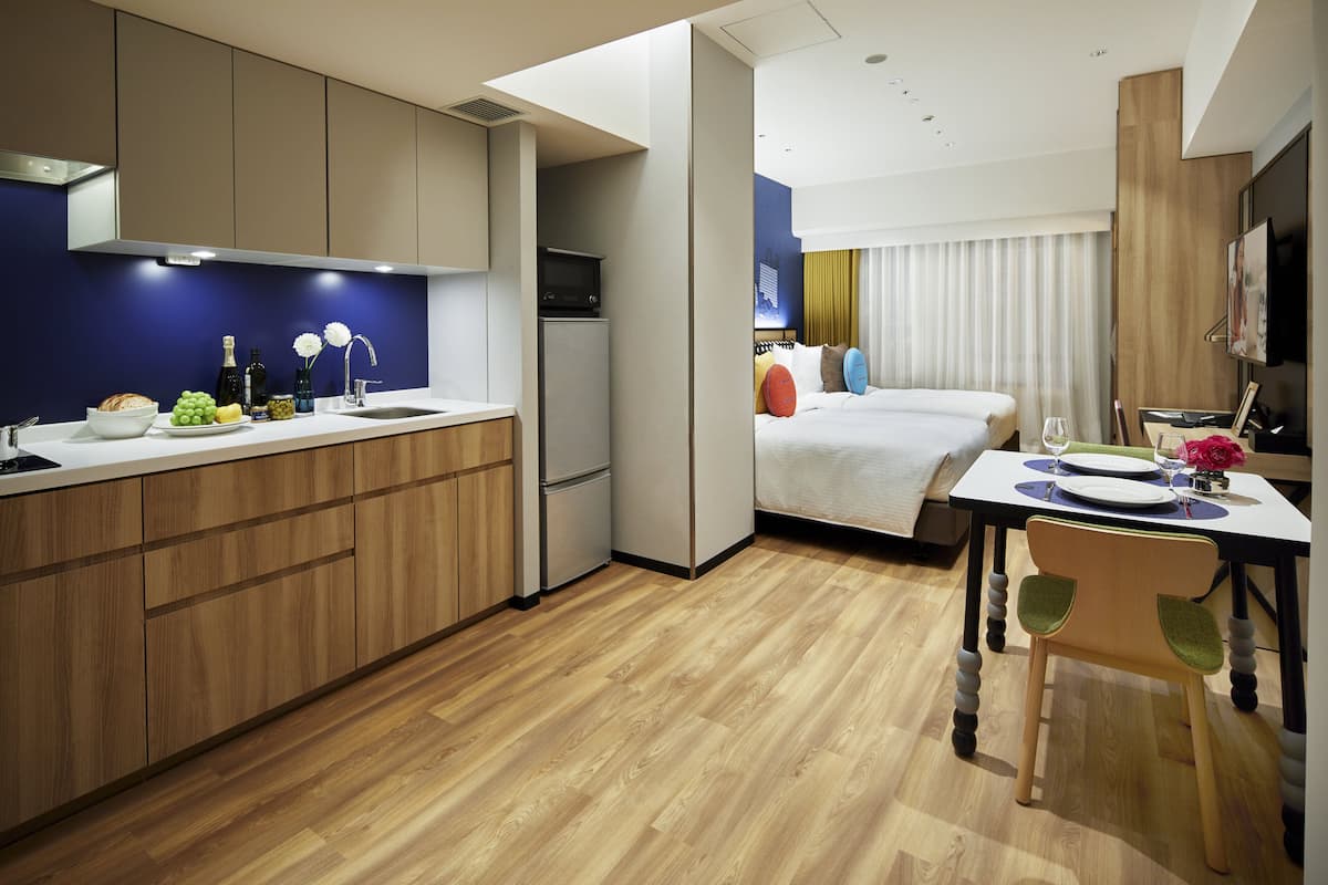 A compact hotel room with a kitchenette, a bed with colorful pillows, a dining area with a table and chair, and a TV mounted on the wall.