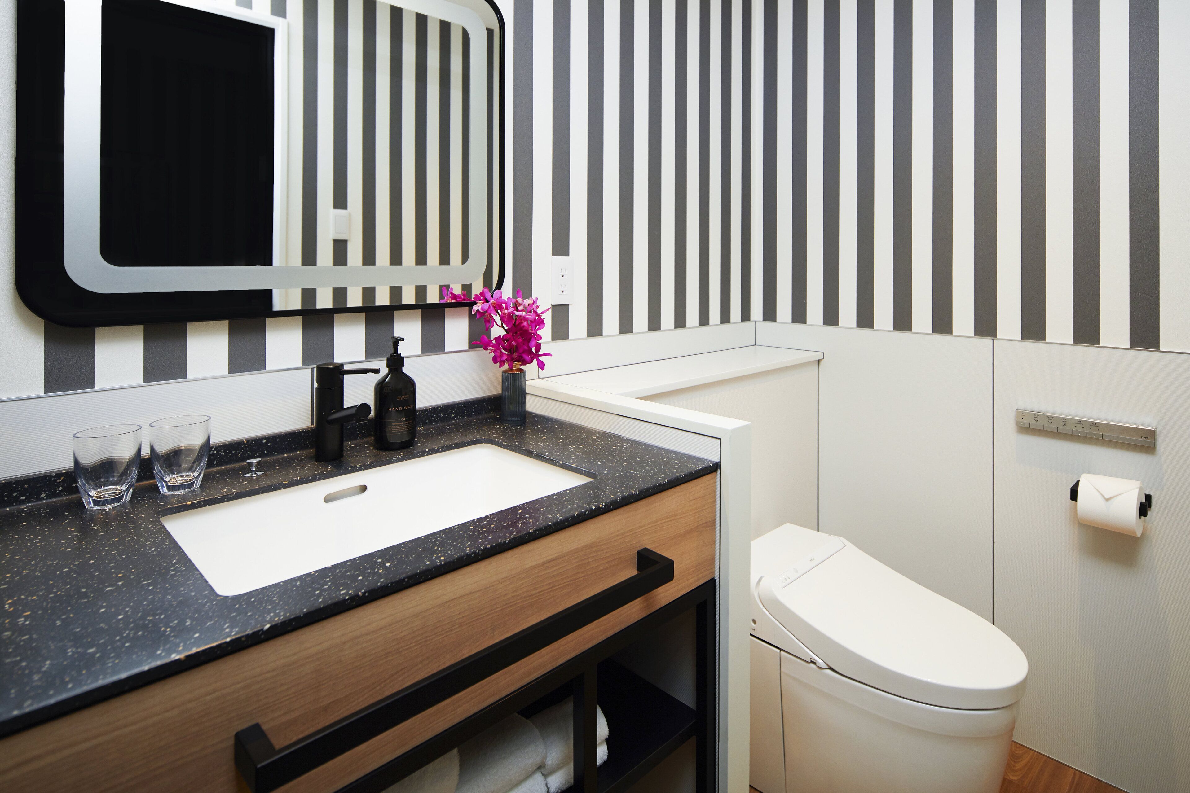 A modern bathroom with a black speckled countertop, a white sink, a black faucet, a mirror, a toilet, and a striped wall.