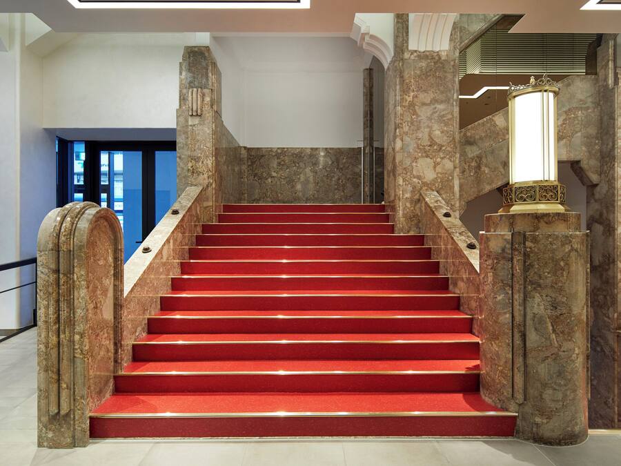 A grand staircase with red carpeted steps, marble walls, and a decorative column with a crown-like top.