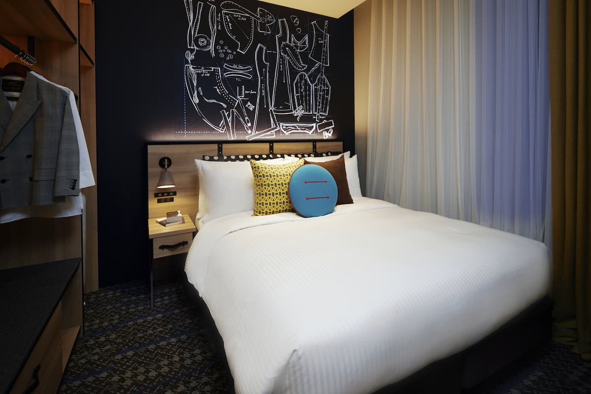 A hotel room with a neatly made bed, a headboard with a light fixture, a bedside table with a lamp, and a wall with a geometric design.