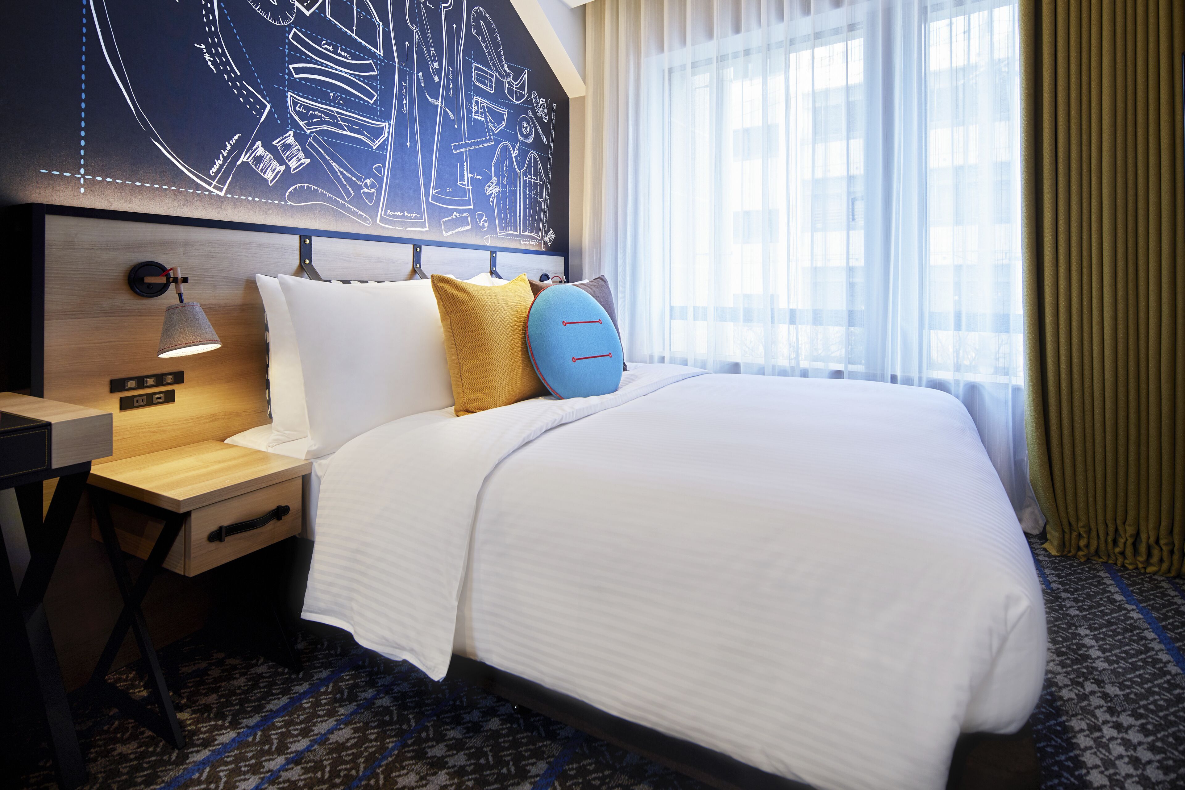 A hotel room with a large bed, a bedside table with a lamp, a wall with a blueprint design, and a window with curtains.