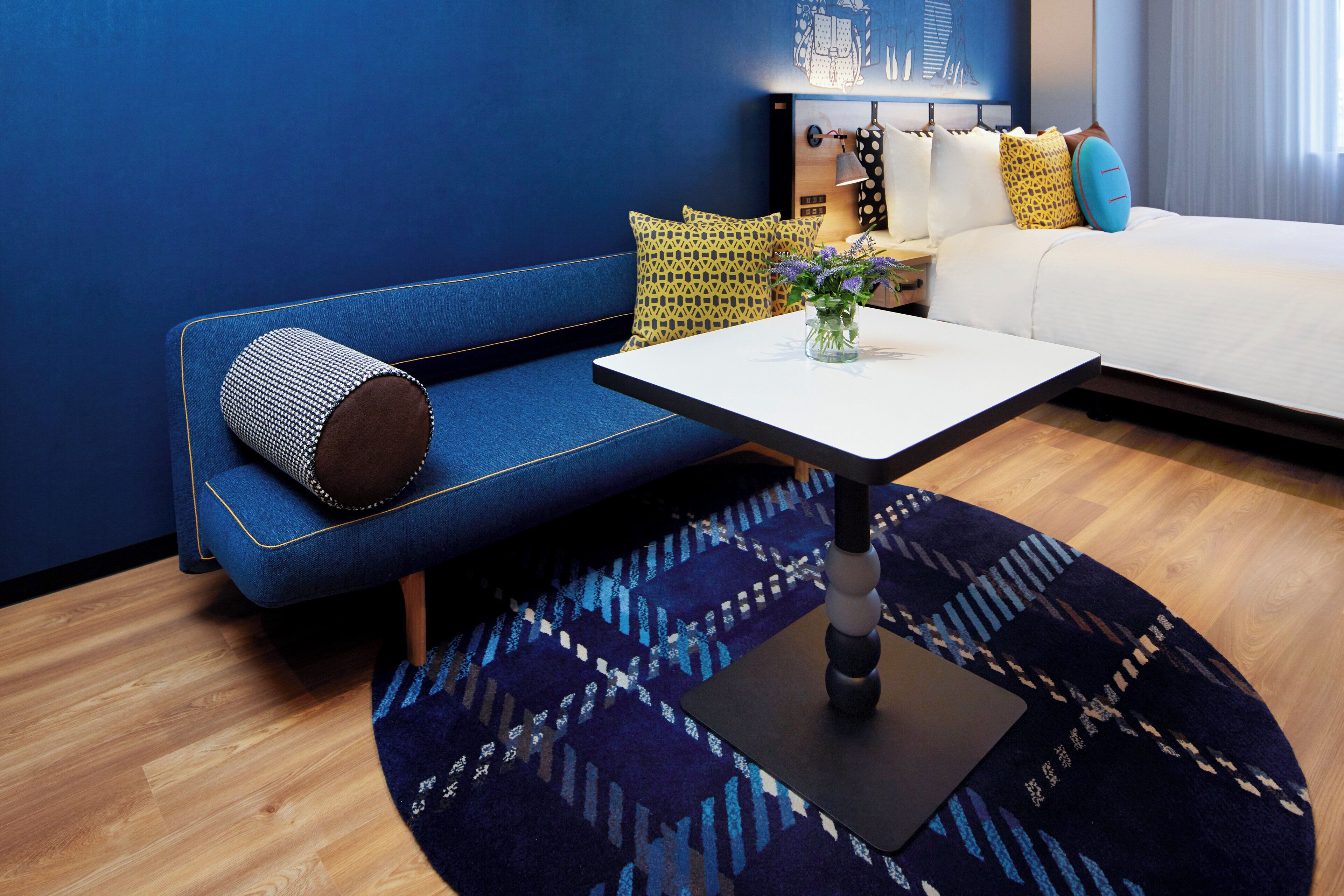 A modern hotel room with a blue sofa, a white bed, a small table, and a patterned rug.