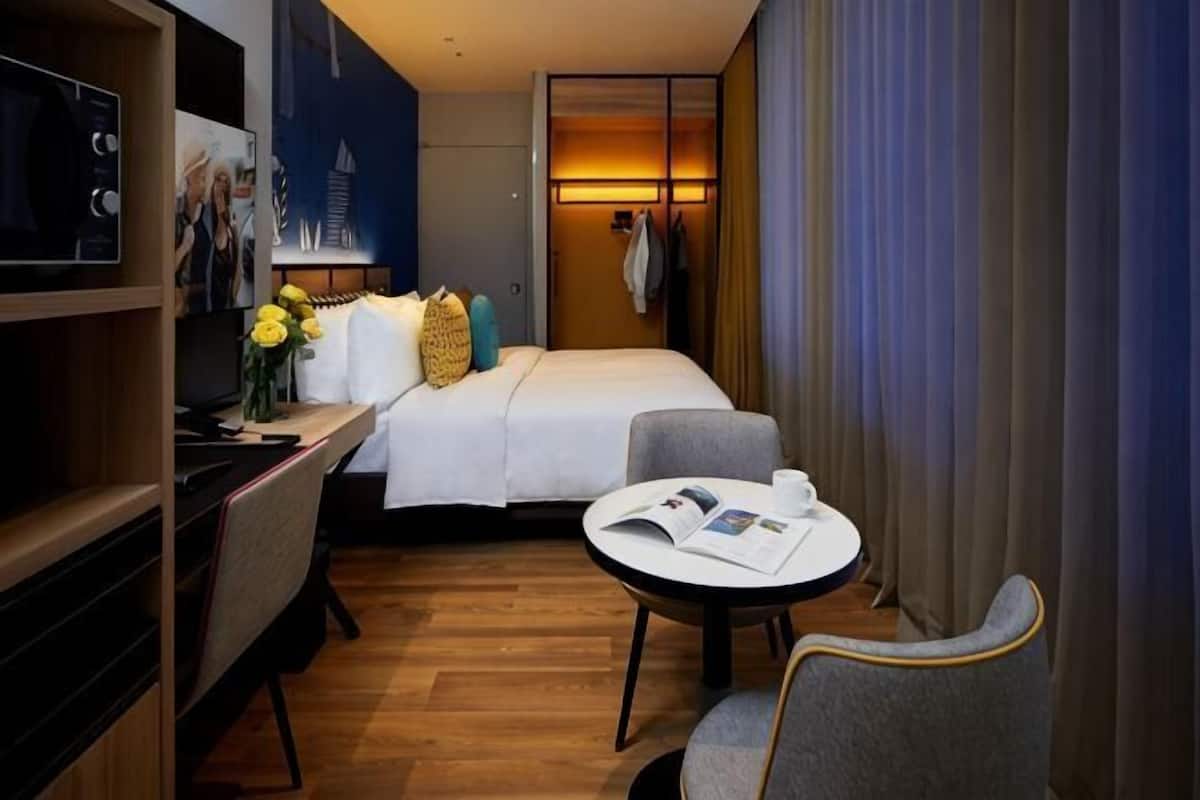 A hotel room with a bed, a desk with a computer, a chair, a small table, and a TV.