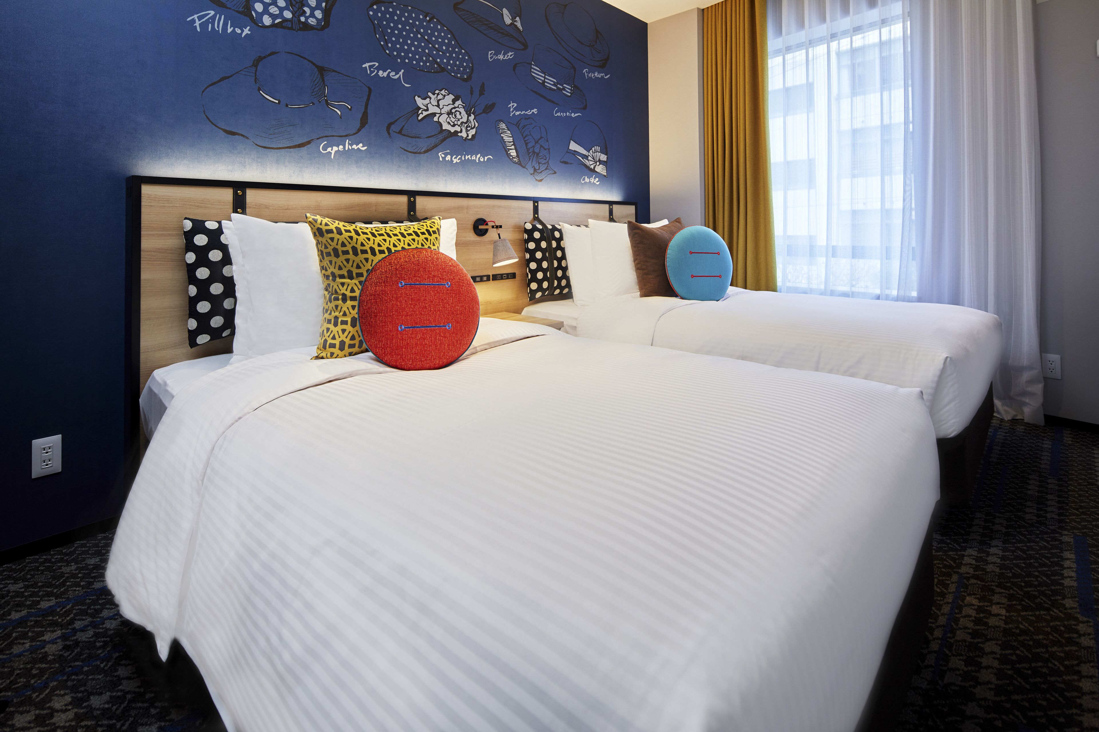 Quarto de hotel com duas camas, parede azul com obras de arte e uma janela com cortinas.