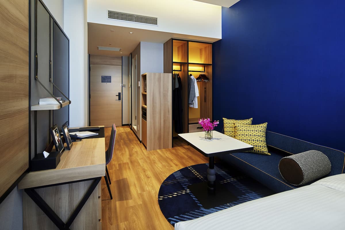 A modern hotel room with a wooden desk, a sofa with cushions, a small table with a vase, and a wardrobe.