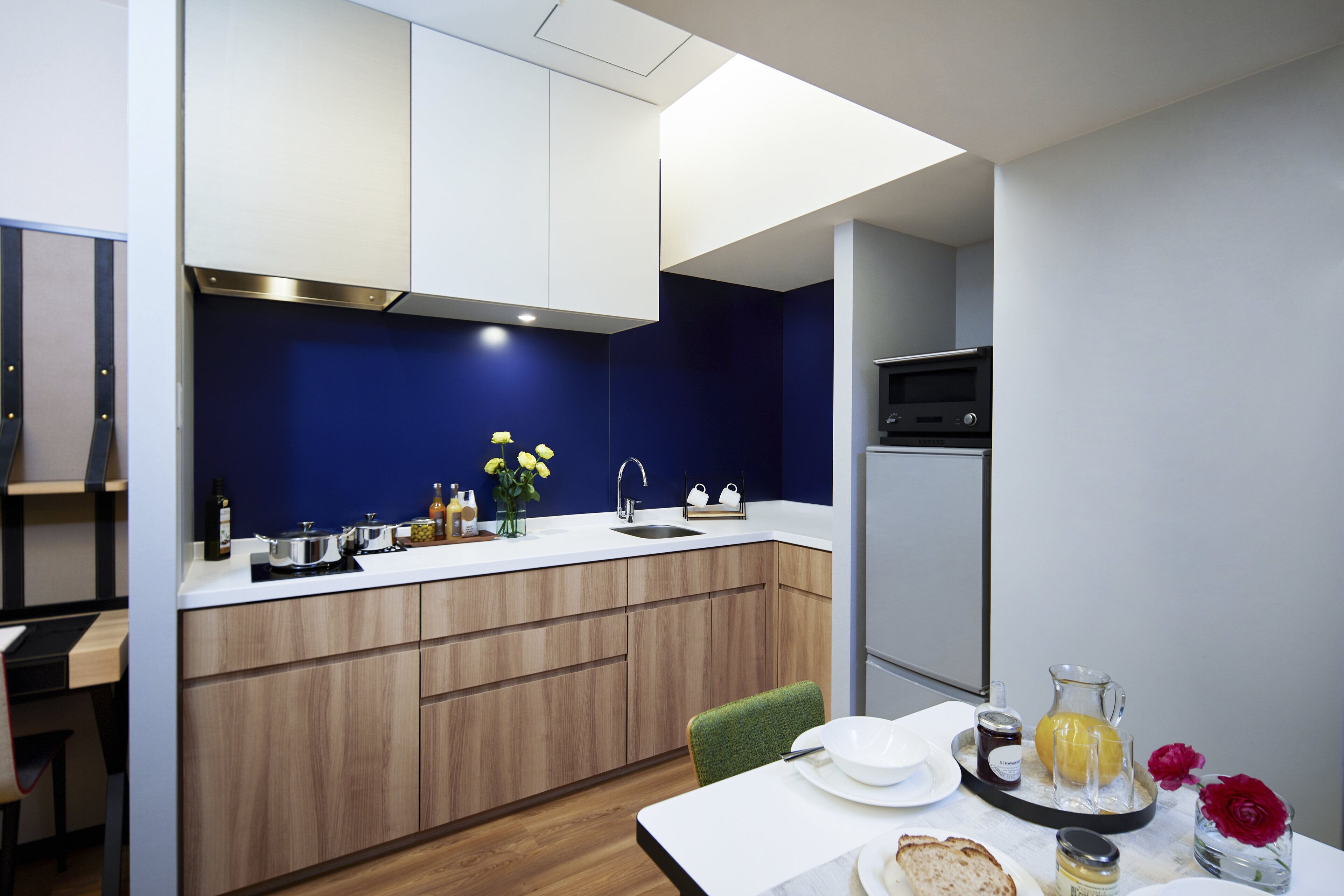 A modern kitchen with wooden cabinets, a white countertop, and a sink. There is a dining table with a green chair and a tray with a pitcher of juice and a vase with flowers.