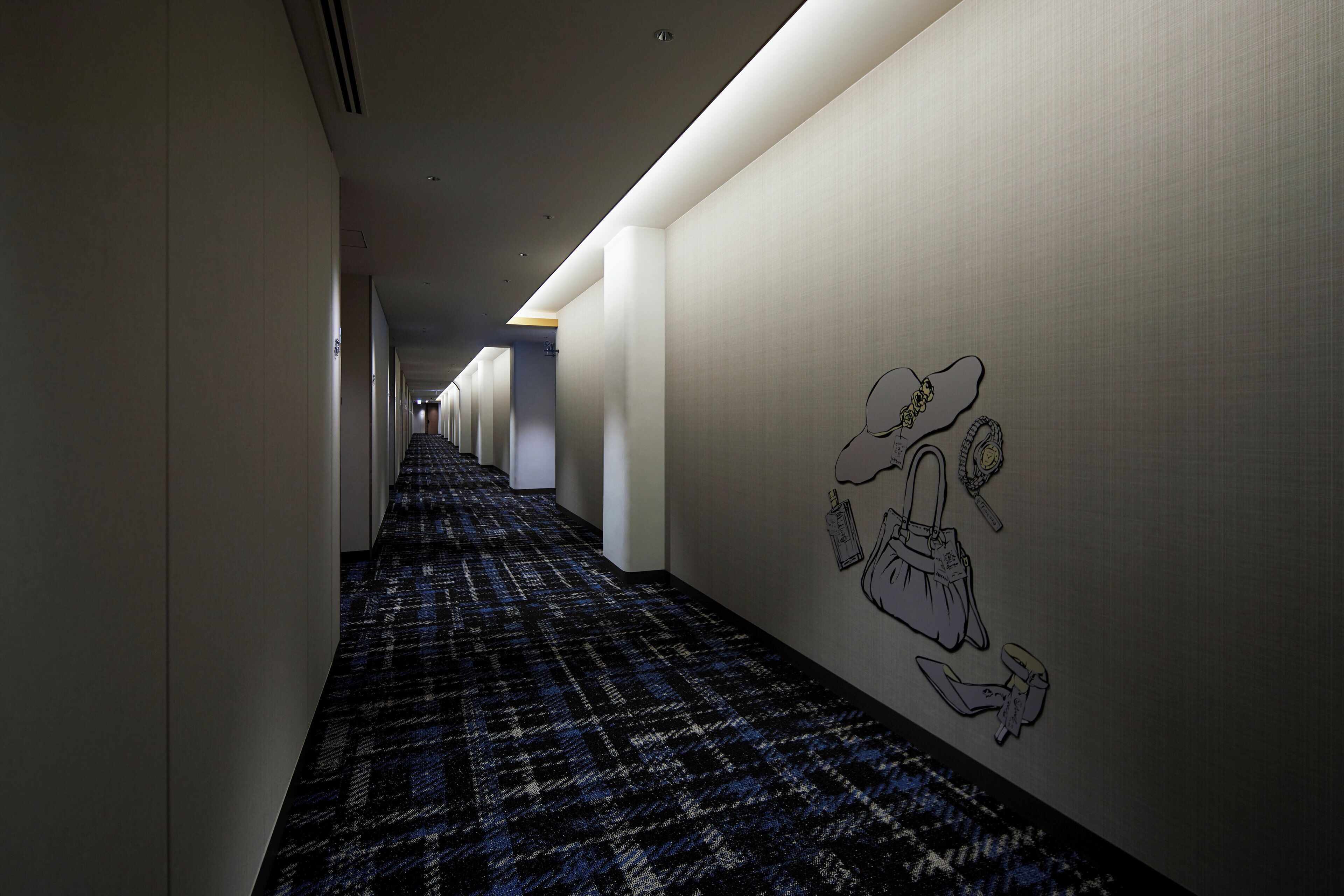 A hotel hallway with a patterned carpet, white walls, and a wall-mounted illustration of a handbag and accessories.
