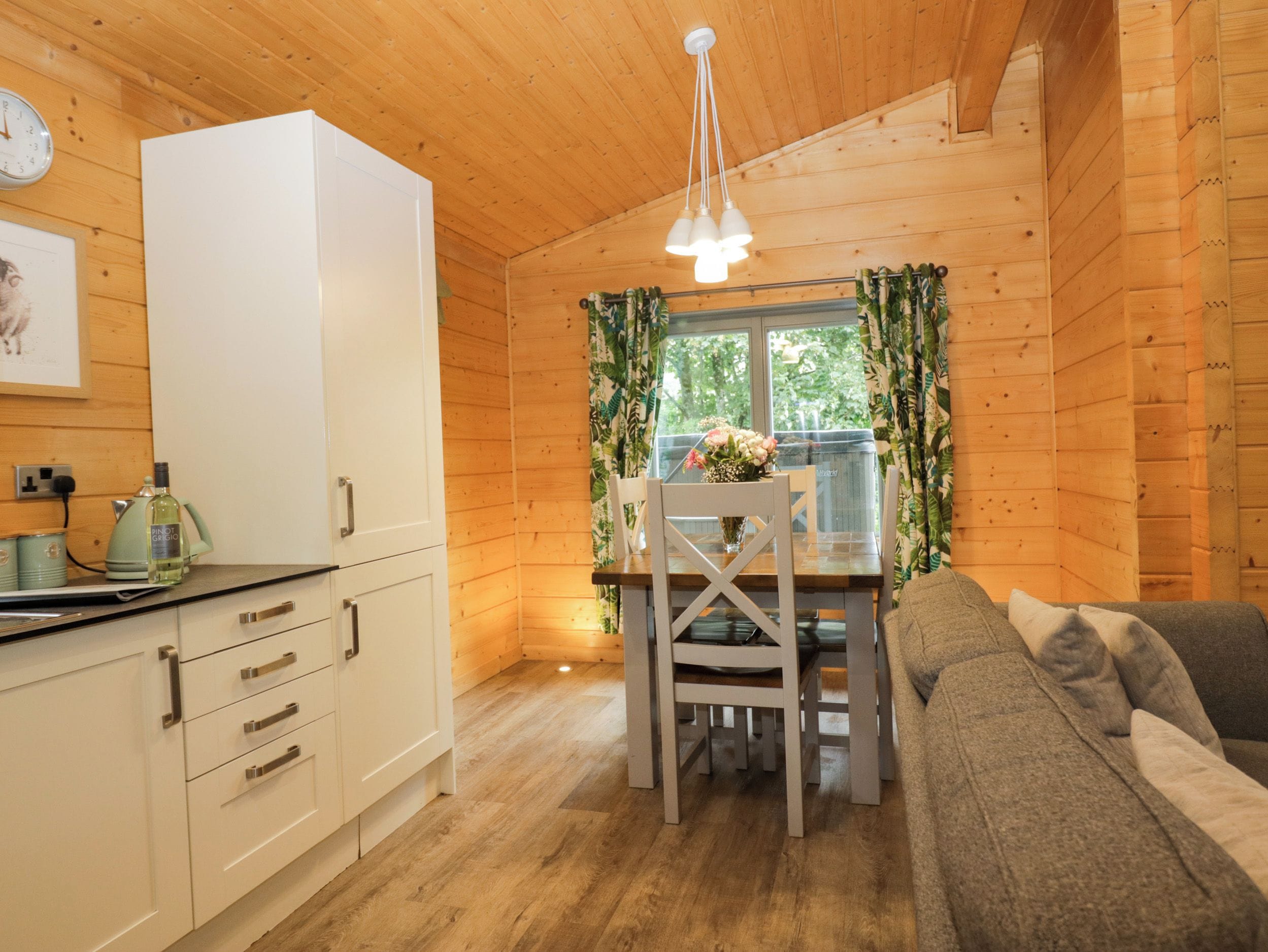 A cozy cabin interior with a dining table, chairs, a sofa, a white refrigerator, and a clock on the wall.