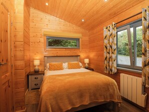 A wooden cabin bedroom with a bed, bedside lamps, a window with curtains, and a view of greenery outside. - Oak Lodge (Ulverston)