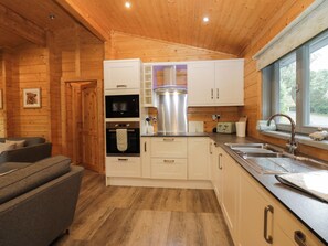 A compact kitchen with wooden cabinets, a built-in oven, a stainless steel range hood, and a window with a view of greenery. - Oak Lodge (Ulverston)
