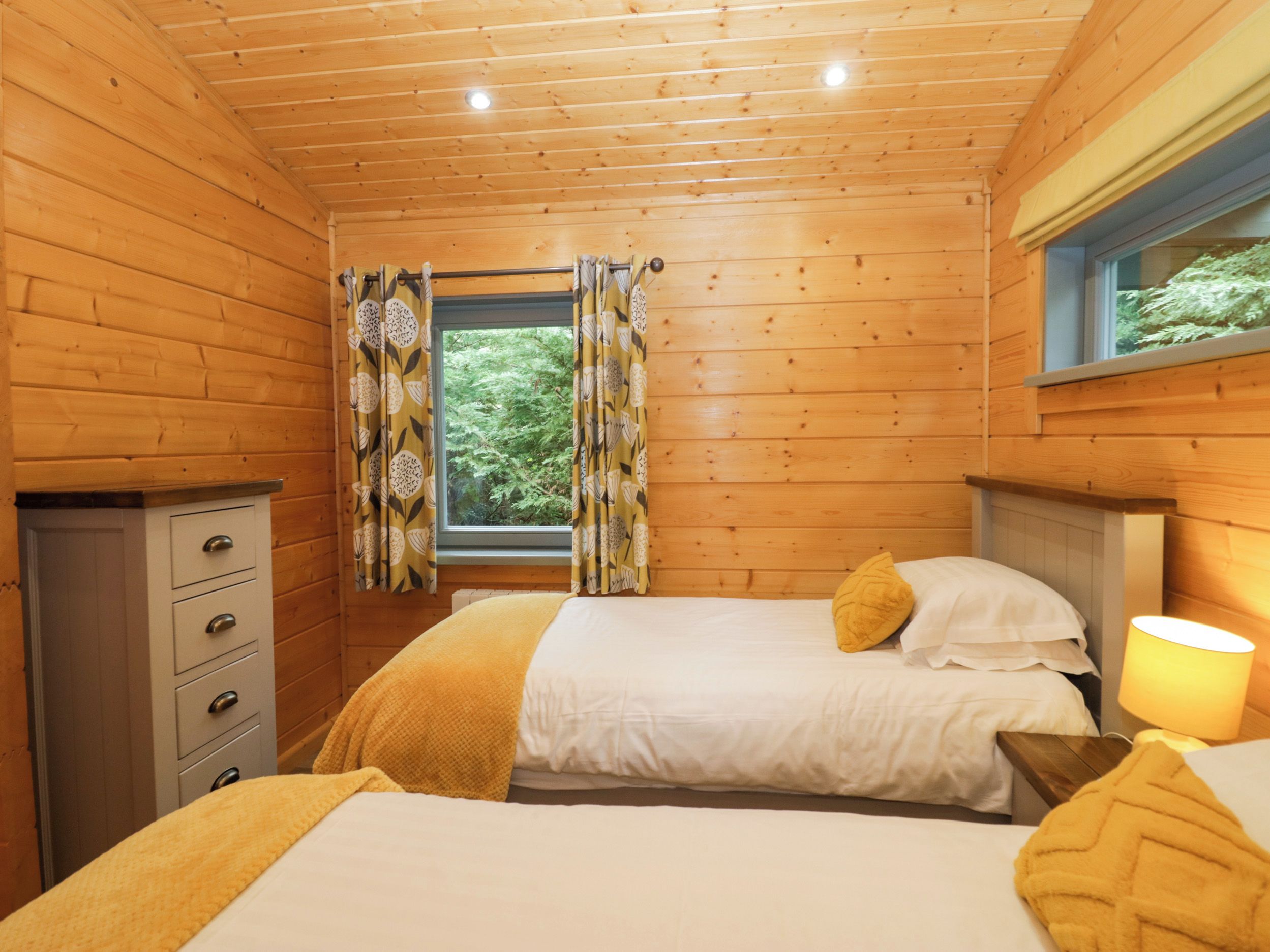 A wooden cabin bedroom with two beds, a dresser, and a window with curtains.