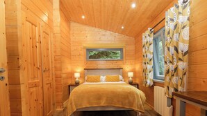 A wooden cabin bedroom with a bed, bedside lamps, a window with curtains, and a radiator. - Oak Lodge (Ulverston)