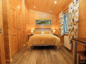 A wooden cabin bedroom with a bed, bedside lamps, a window with curtains, and a radiator. - Oak Lodge (Ulverston)