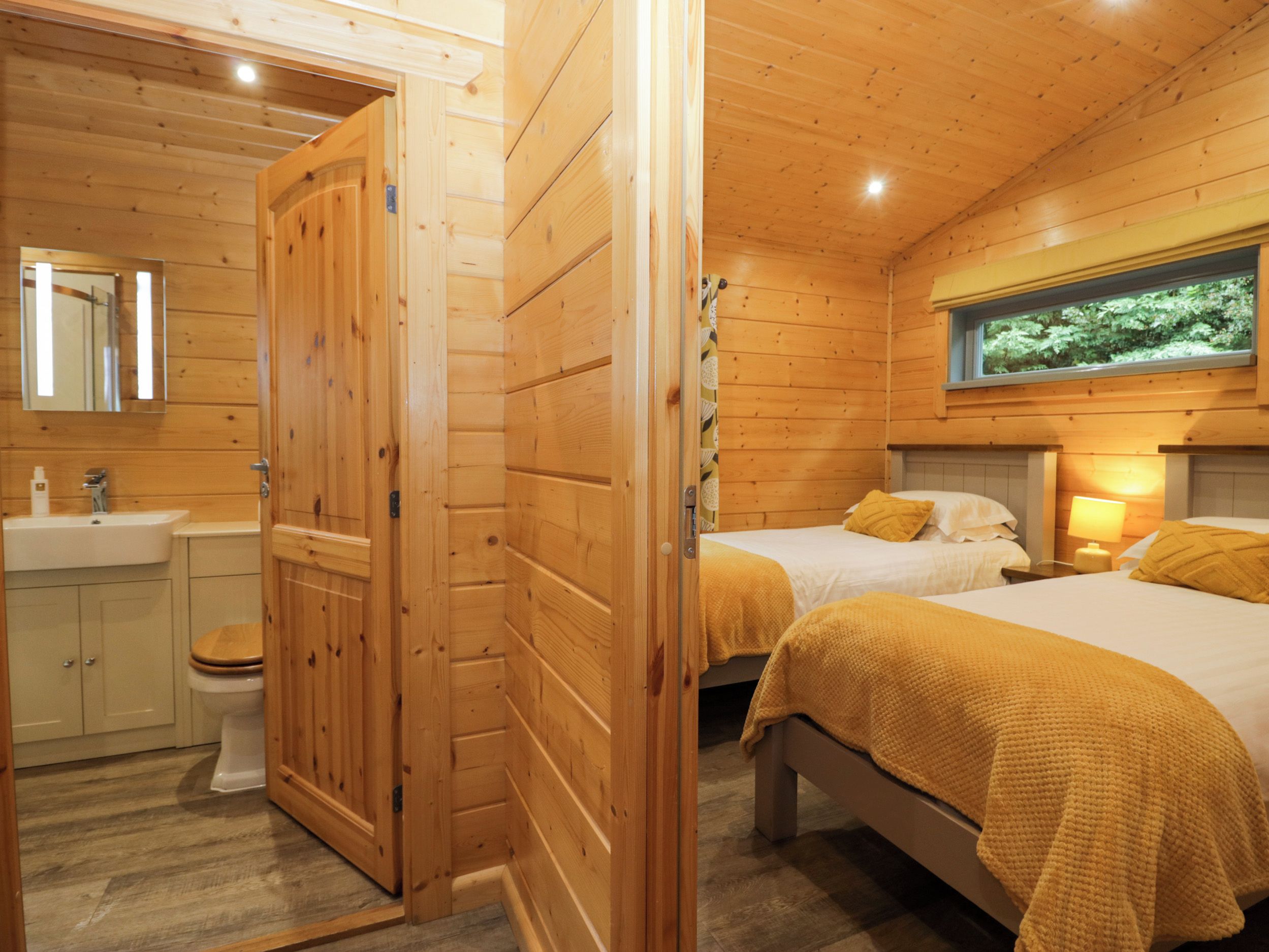 Una cabaña de madera con baño, dos camas con ropa de cama amarilla y un ventanal con vistas al follaje.