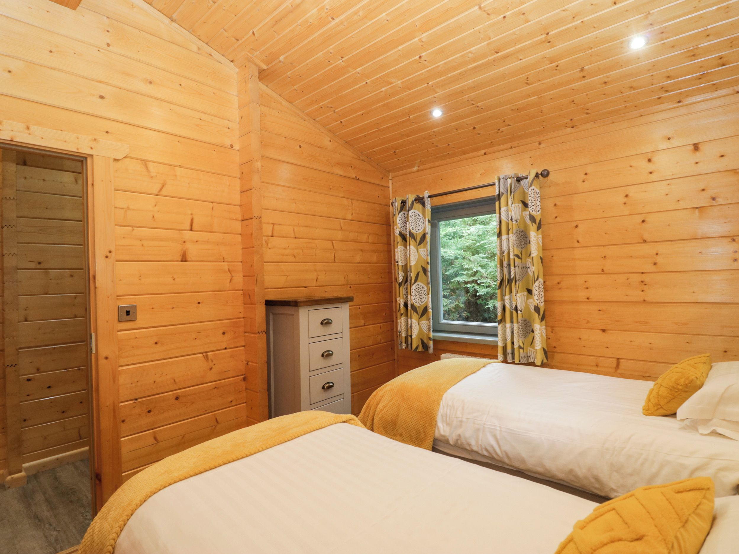 A wooden cabin room with two beds, a small cabinet, and a window with curtains.