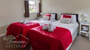A hotel room with two beds, each with red and white bedding, a window with curtains, a bedside table with a lamp, and a small bookshelf. - Redcliff View Lodge (Weymouth)