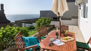 A wooden table with a bottle, glasses, and a newspaper, set under a beige umbrella on a patio with a view of the sea.
