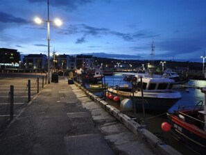 Un puerto deportivo al anochecer con barcos amarrados, un paseo pavimentado y edificios al fondo.