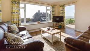 A cozy living room with a brown leather sofa, a white coffee table, and a flat-screen TV on a stand. Large windows with curtains offer a view of the outside.