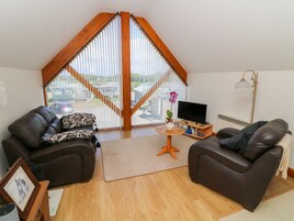 A cozy living room with a sloped ceiling, two leather sofas, a small coffee table, a TV, and a large window with blinds.