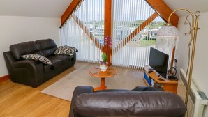 Un salon chaleureux avec un canapé en cuir marron, un divan en cuir noir et une table basse en bois. Un téléviseur est posé sur un meuble en bois, et une lampe se trouve sur la droite.