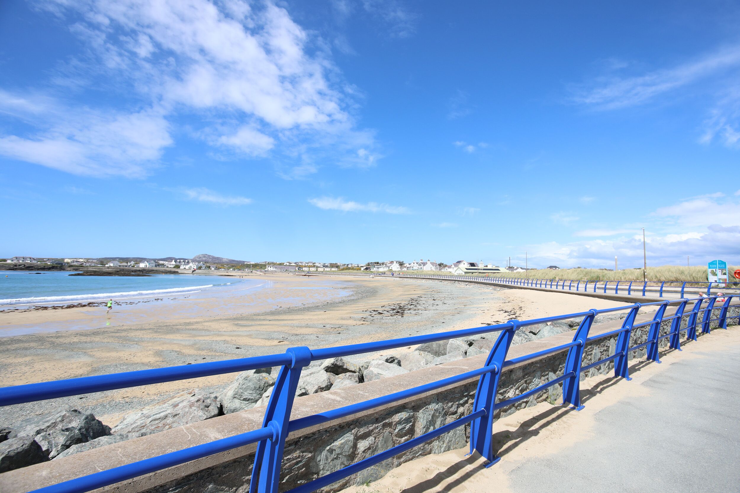 Un paseo marítimo junto a la costa, con barandilla azul, que conduce a una playa de arena y un litoral rocoso.