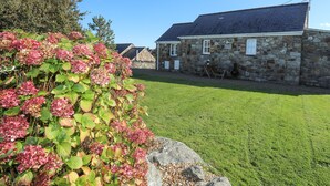 A stone building with a sloped roof, a grassy yard, and a large bush with pink flowers. - Tyn Towyn - Bwthyn Carreg (Holyhead)