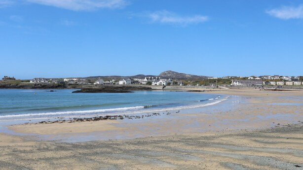 A coastal beach with a clear sky, sandy shore, and houses in the background. - Tyn Towyn - Bwthyn Carreg (Holyhead)
