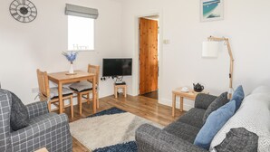 A living room with a sofa, a TV, a dining table, and a window. - Tyn Towyn - Bwthyn Carreg (Holyhead)