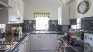 A modern kitchen with white cabinets, a black countertop, a stainless steel stove, and a window with a polka dot valance. - Tyn Towyn - Bwthyn Carreg (Holyhead)