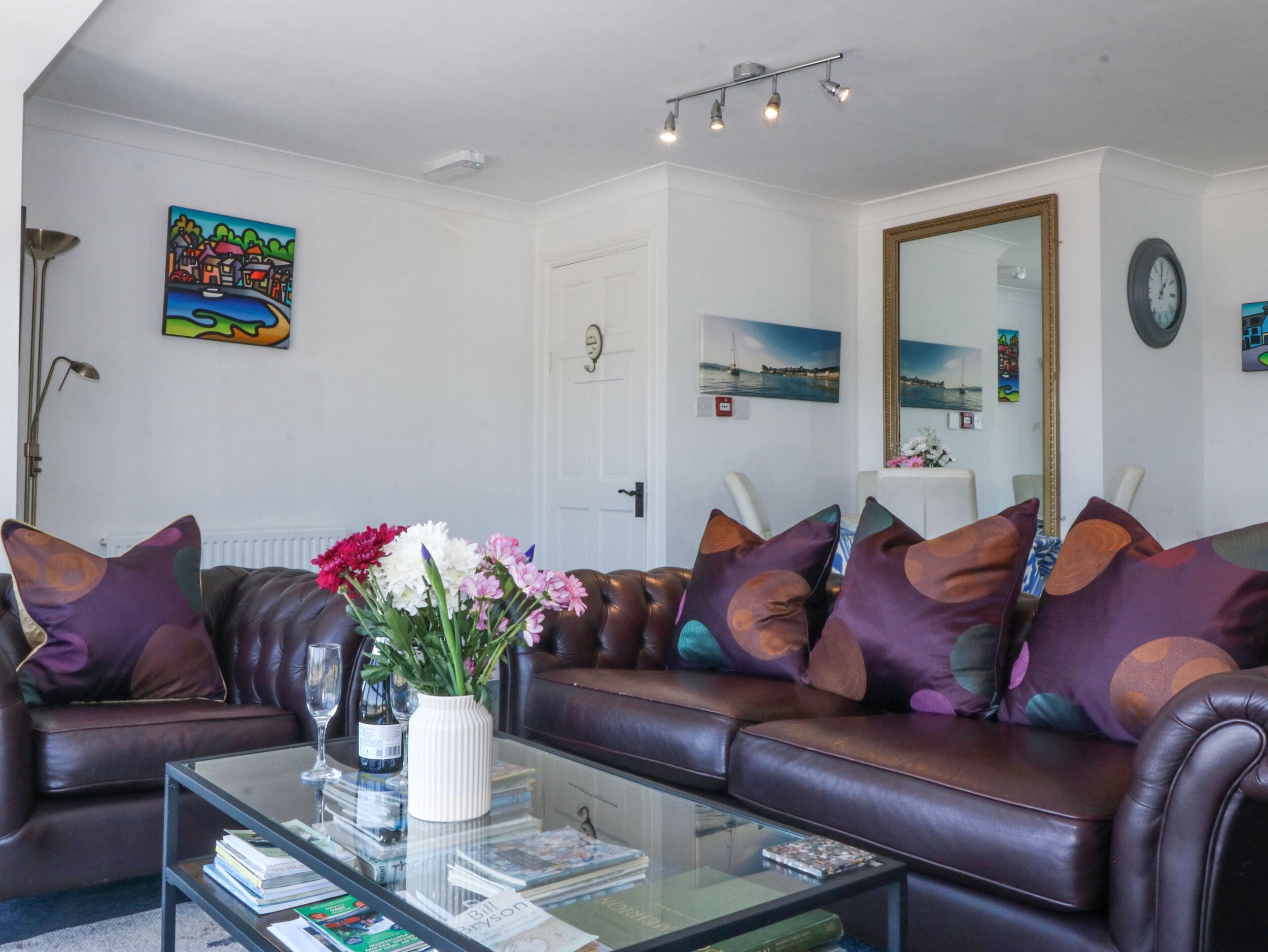 A living room with a brown leather sofa, a glass coffee table, and a vase with flowers. There are books on the table, and a painting is on the wall.
