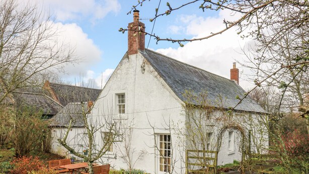 Une maison blanche avec un toit à forte pente, une cheminée et un petit jardin agrémenté d’un banc en bois et d’un sentier.