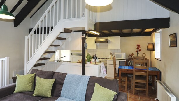 Ein gemütliches Wohnzimmer mit braunem Sofa, grünen Kissen, einer Treppe und einem Küchenbereich.