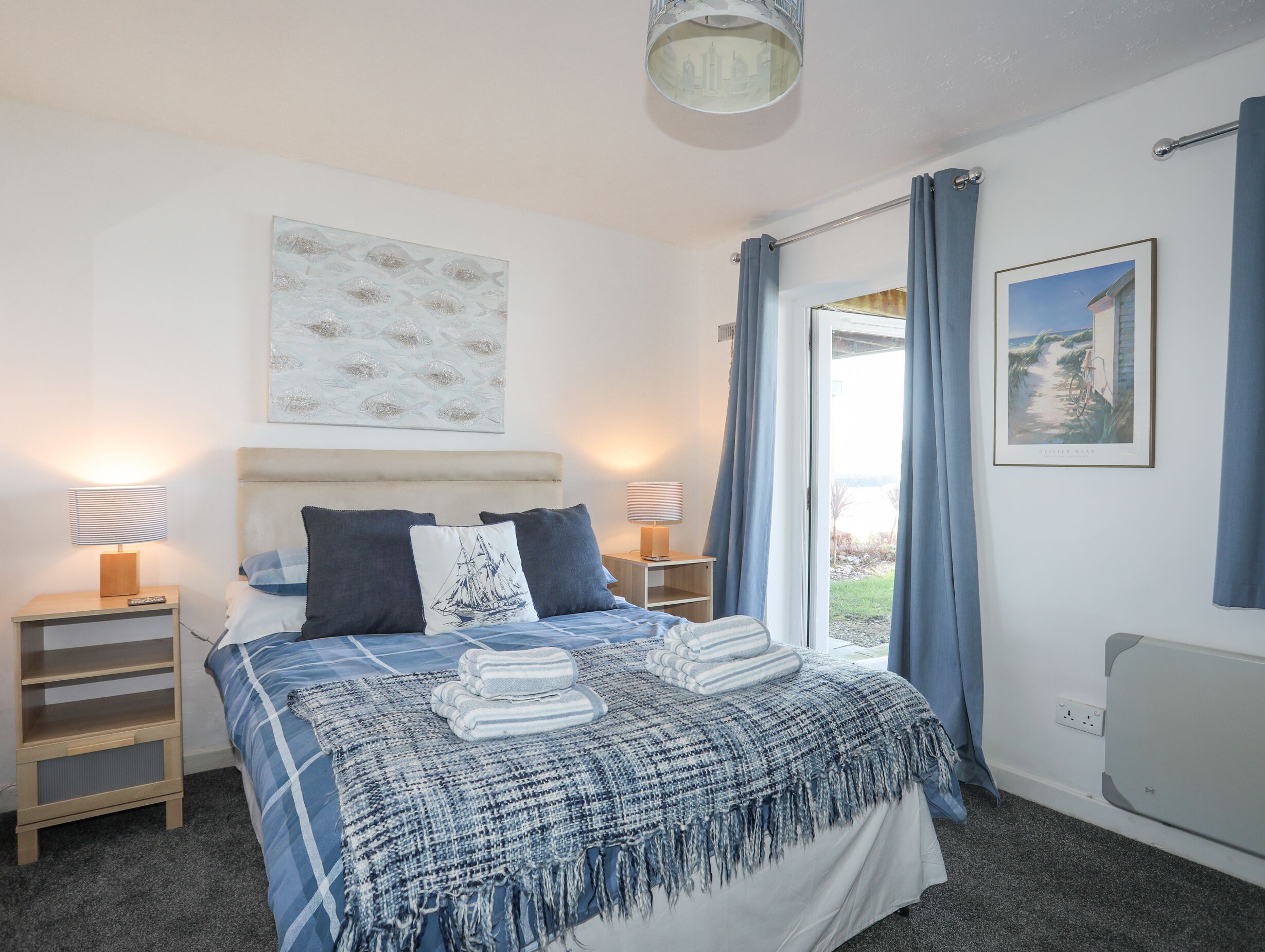 A bedroom with a bed, two bedside tables, a nightstand, a window with curtains, a wall-mounted lamp, and a framed picture.
