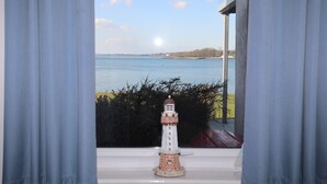 Vista a través de una ventana con cortinas, que muestra un pequeño faro de modelo en un alféizar, una extensión de agua y una costa lejana.