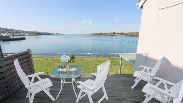 Un balcón con vistas a un lago, sillas blancas y una mesa con un florero.