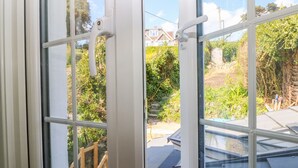 Blick durch ein Fenster mit weißem Rahmen auf einen Garten mit einer Holzterrasse und einem Haus im Hintergrund.