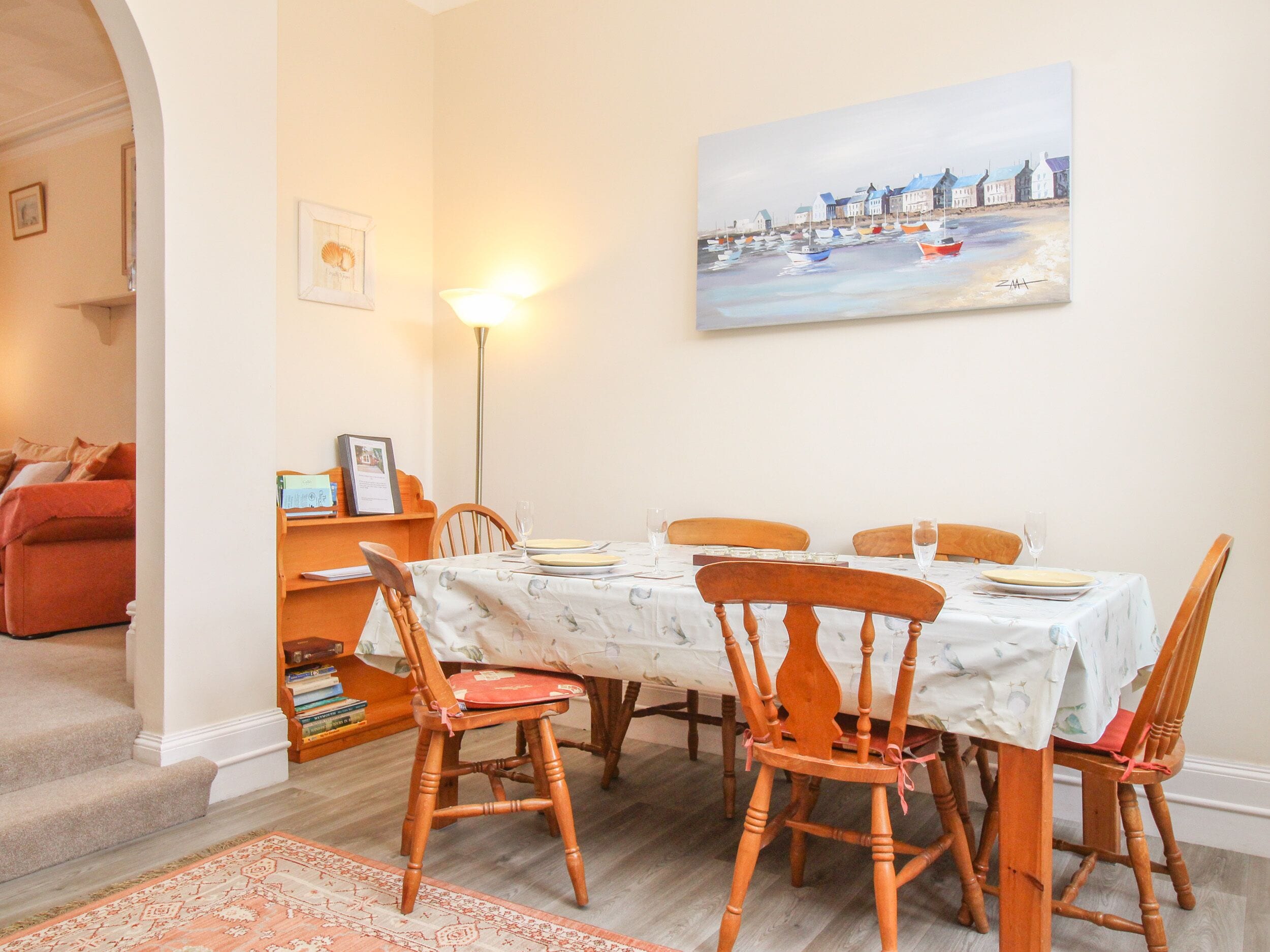 A dining area with a table set for four, surrounded by wooden chairs, a patterned rug, and a painting of a coastal scene on the wall.