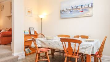 A dining area with a table set for four, surrounded by wooden chairs, a patterned rug, and a painting of a coastal scene on the wall.