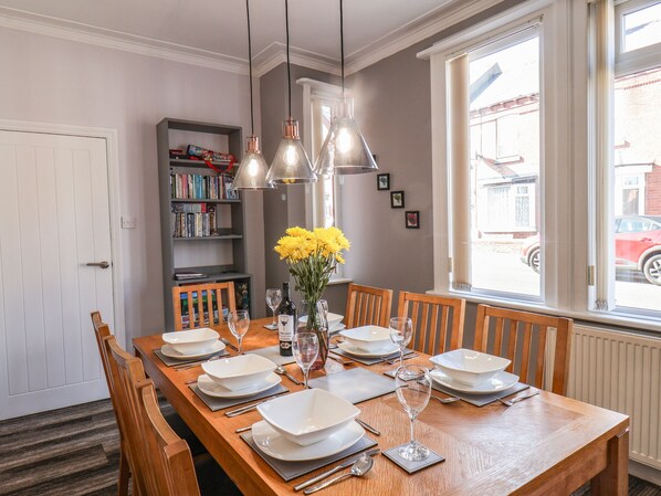 Un coin repas avec une table en bois dressée pour six personnes, entourée de chaises. Des étagères à livres sont adossées au mur, il y a une fenêtre donnant sur la rue et un vase rempli de fleurs jaunes sur la table.