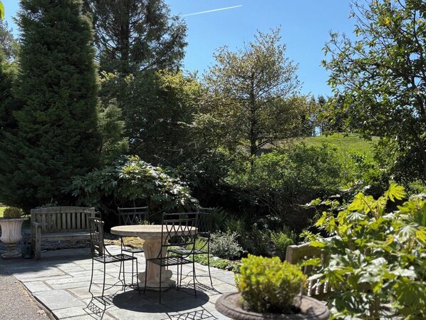 Een terras met een ronde tafel en stoelen, omgeven door weelderige begroeiing en bomen.