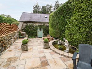 Een terras van natuursteen met een houten tafel en stoelen, een kleine fontein en een groene deur.