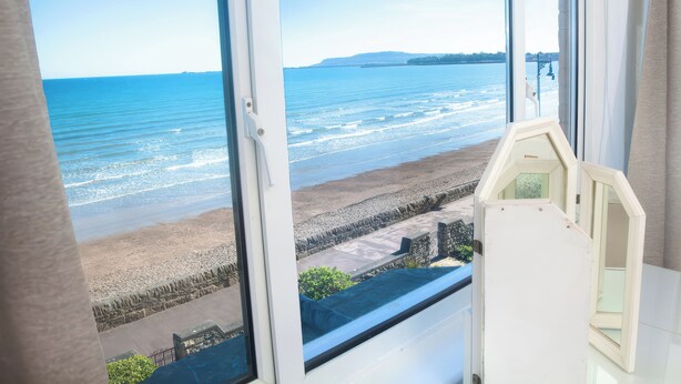 Een kamer met een groot raam met uitzicht op een strand, een stenen muur en een kleine witte tafel.