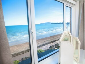 A room with a large window overlooking a beach, a stone wall, and a small white table. - Weymouth Bay Apartment C (Weymouth)