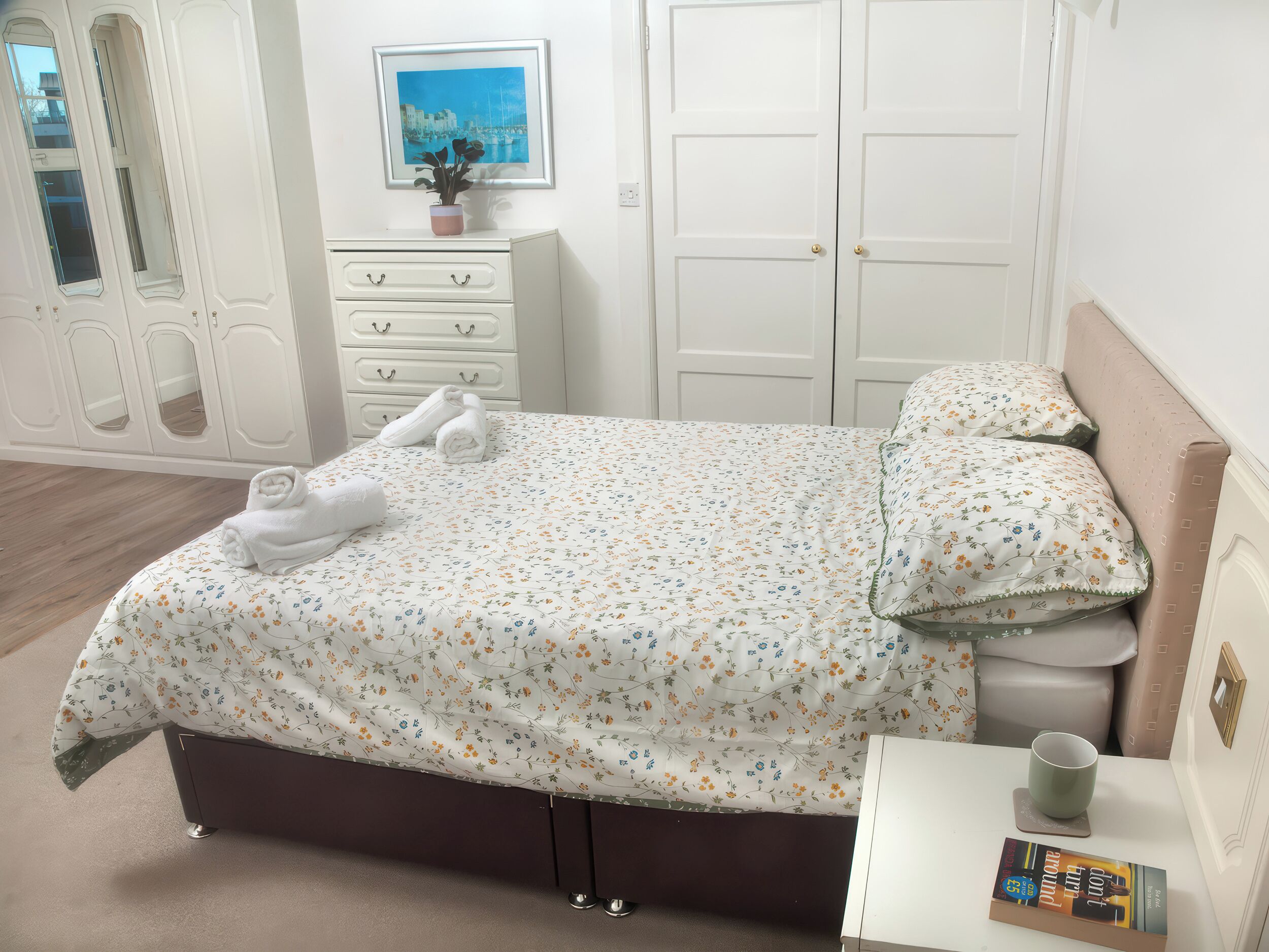 A neatly made bed with a floral bedspread, a bedside table with a book and a cup, and a dresser with a mirror.