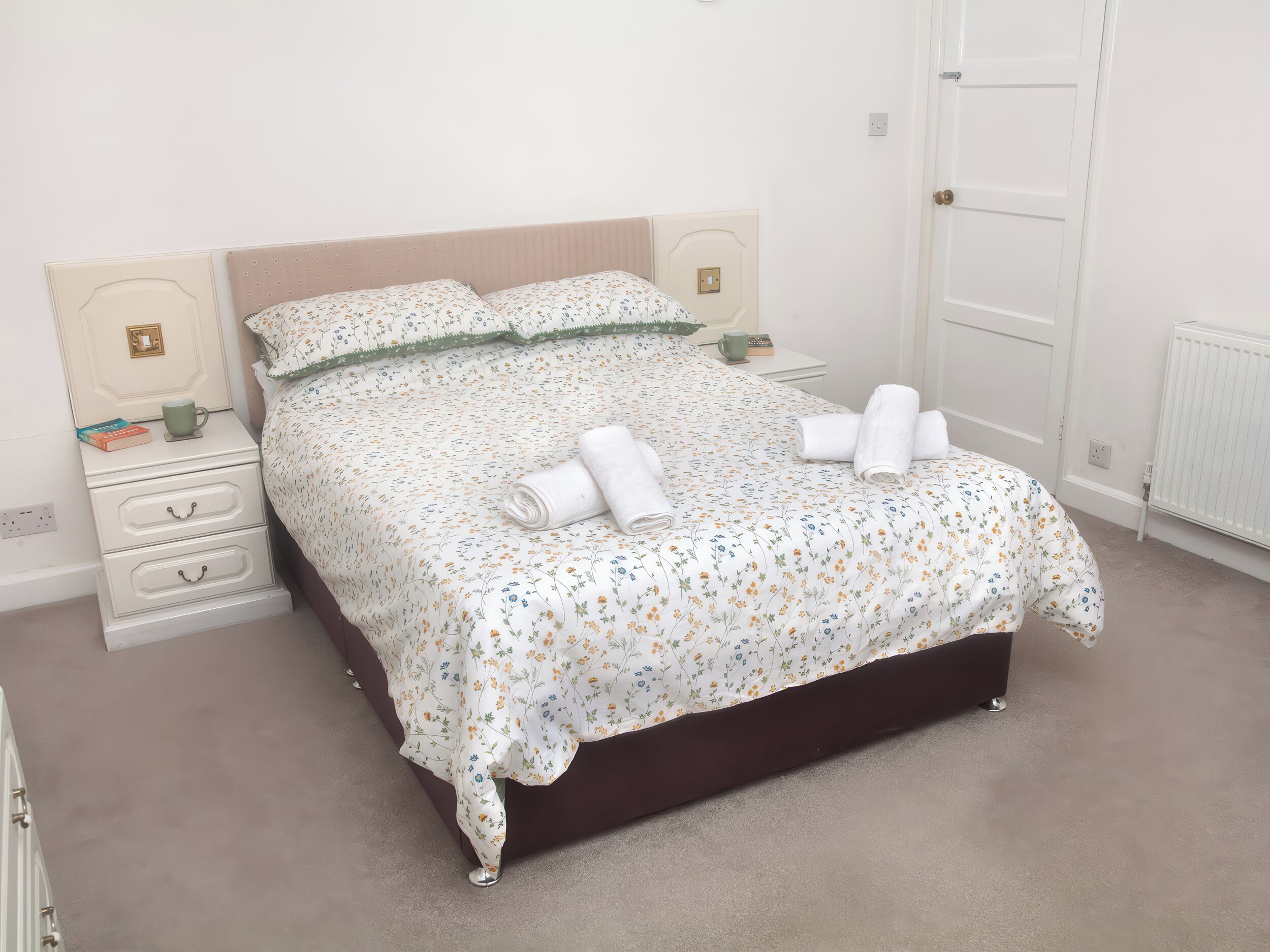 Una cama bien hecha con ropa de cama floral, una mesita de noche con un libro y una taza, y una puerta con pomo.