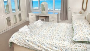 Een slaapkamer met een bed, een kaptafel, een dressoir en een raam met uitzicht op zee.