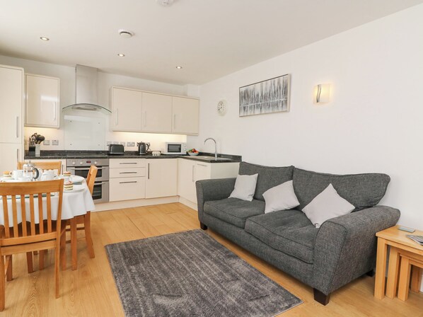 A modern kitchen with white cabinets, a dining table with chairs, and a grey sofa with cushions.