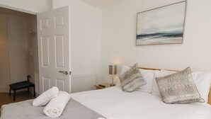 Ein ordentlich bezogenes Bett mit weißen Leinentüchern, einer grauen Tagesdecke und zusammengerollten Handtüchern. An der Wand hängt ein Meeresblick-Gemälde, eine Nachttischlampe und ein kleiner Tisch mit einer Fernbedienung.